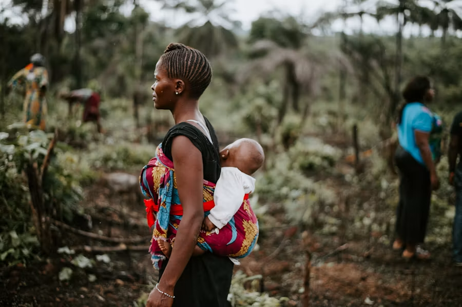 Une femme africaine portant son bébé dans le dos au milieu d'une végétation tropicale, illustrant la résilience et la force des femmes soutenues par SFEM.